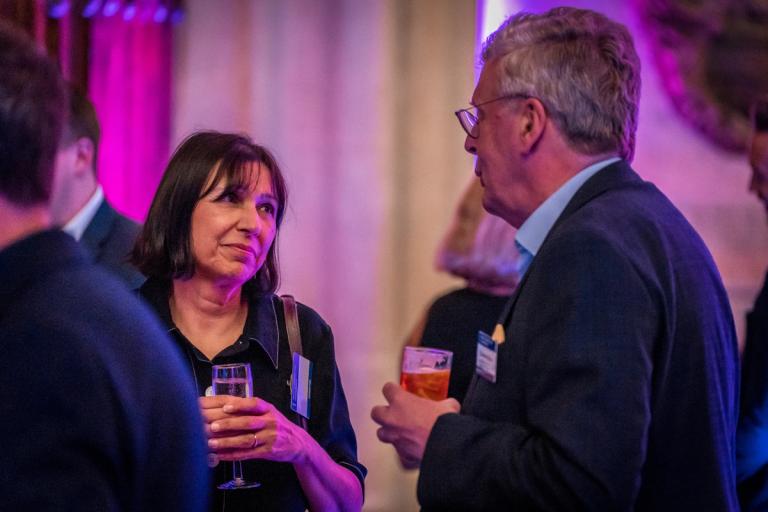 A woman and a man chat, holding drinks, at a social event.