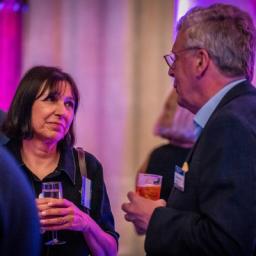 A woman and a man chat, holding drinks, at a social event.