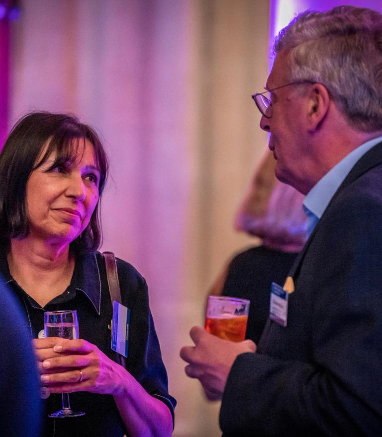 A woman and a man chat, holding drinks, at a social event.