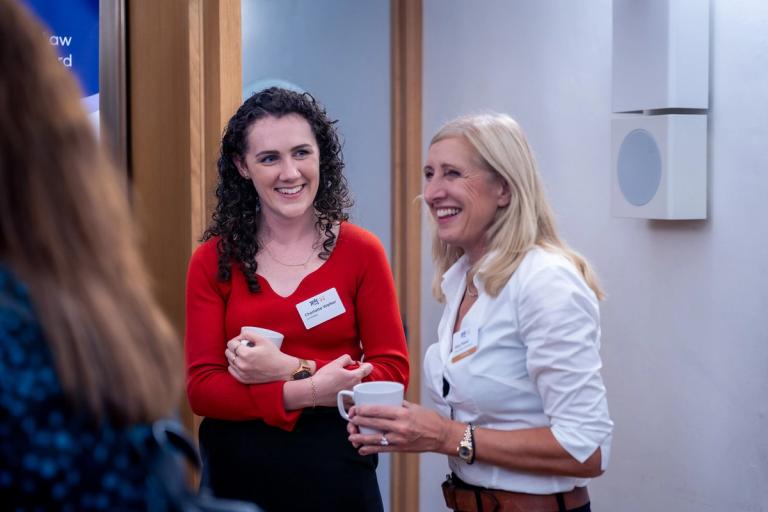 Two smiling women, one in a red top and one in a white shirt, holding mugs.