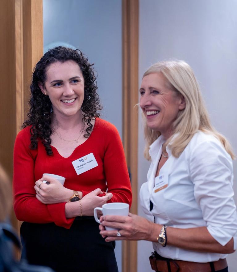 Two smiling women, one in a red top and one in a white shirt, holding mugs.