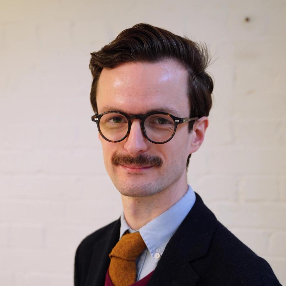 A man with dark hair, glasses, and a mustache, wearing a jacket and yellow tie, smiling.