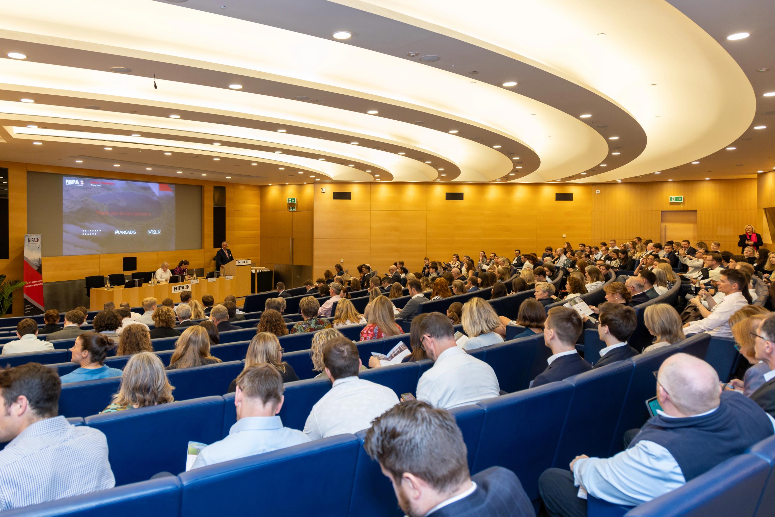A large audience in a modern auditorium, facing a stage with a screen displaying a presentation.
