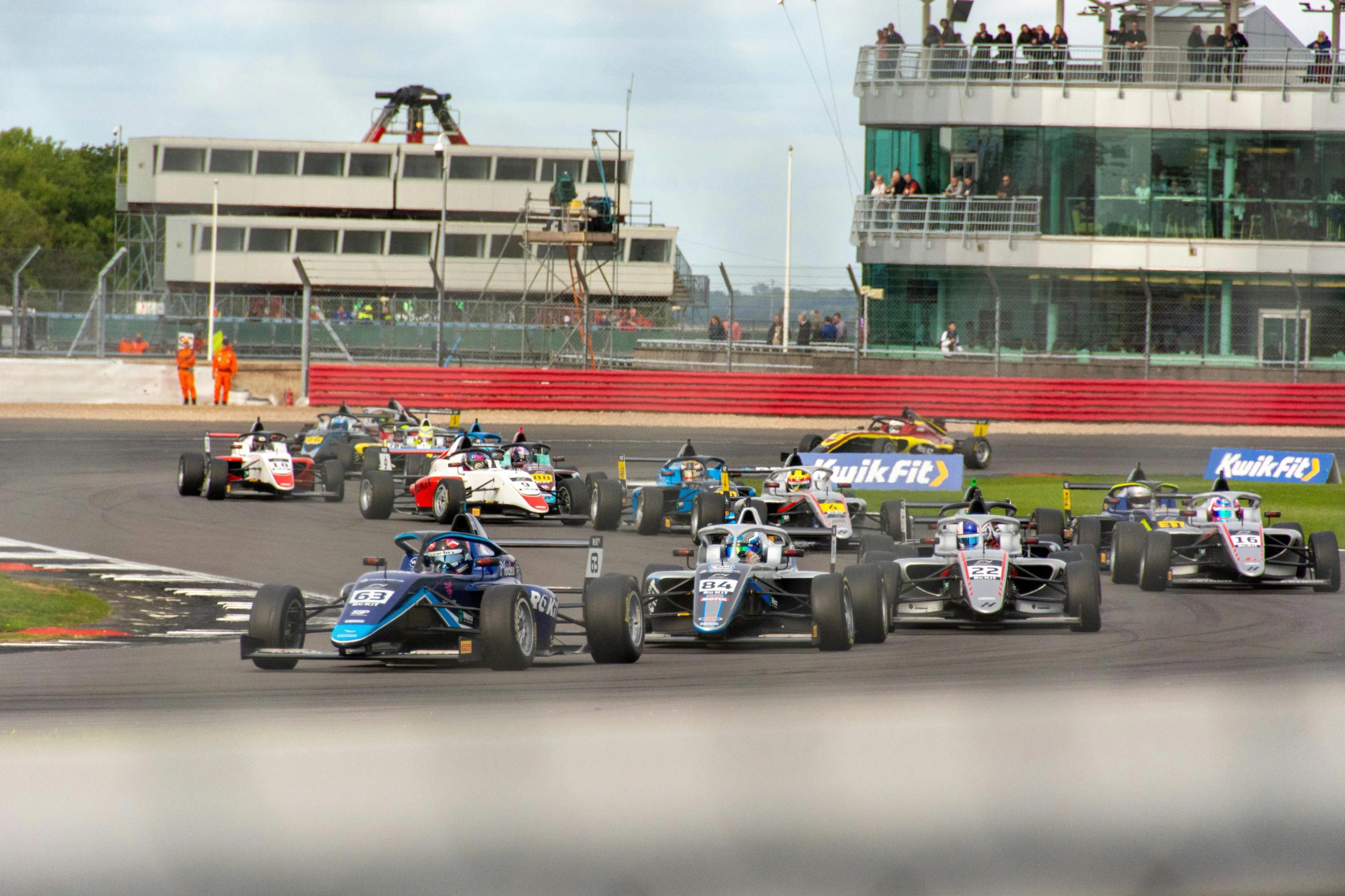 A group of open-wheel race cars on a track, with a blue car in the foreground, rounding a corner past buildings and spectators.