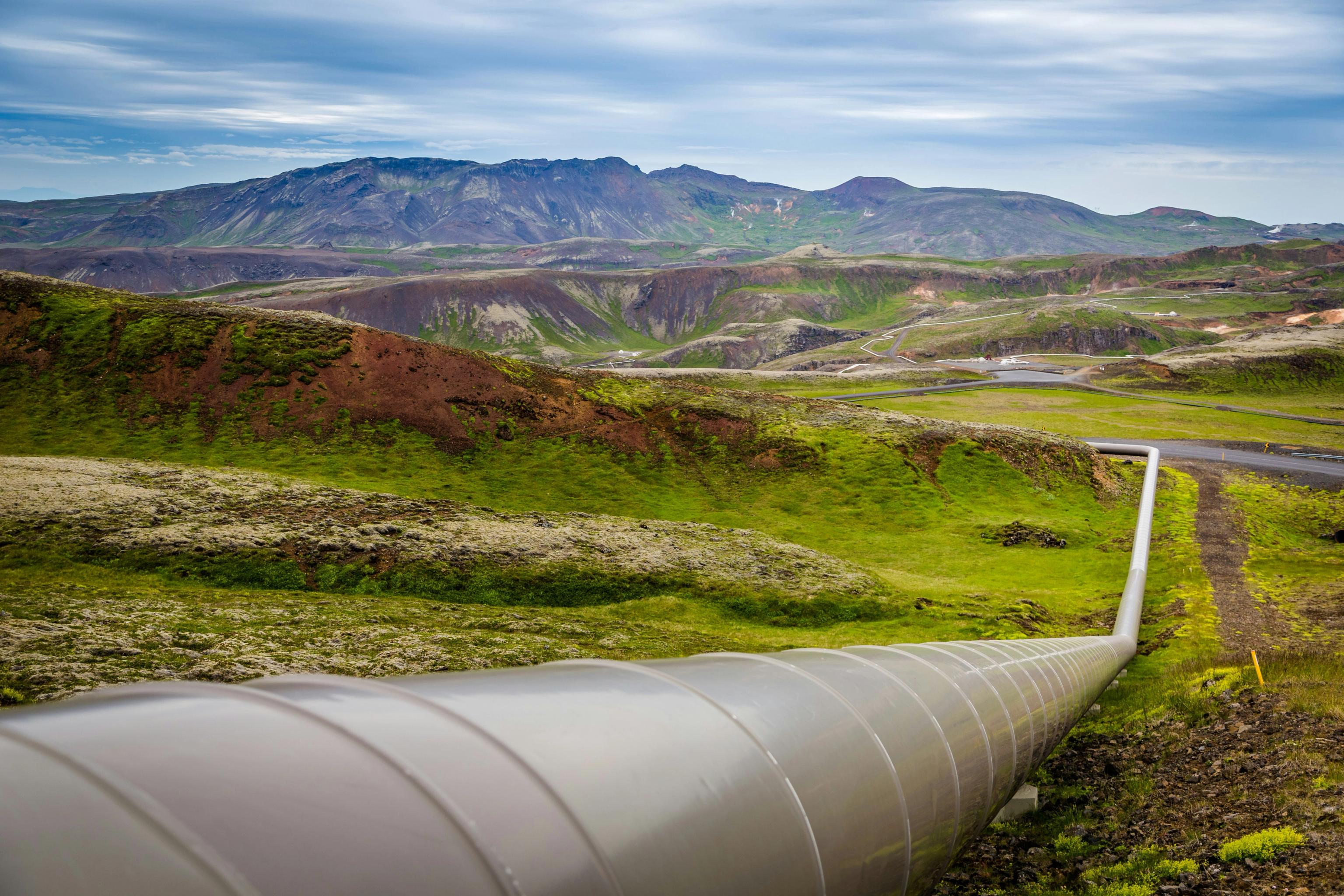 A grey pipeline stretches across a green, mossy volcanic landscape with distant mountains under a cloudy sky.