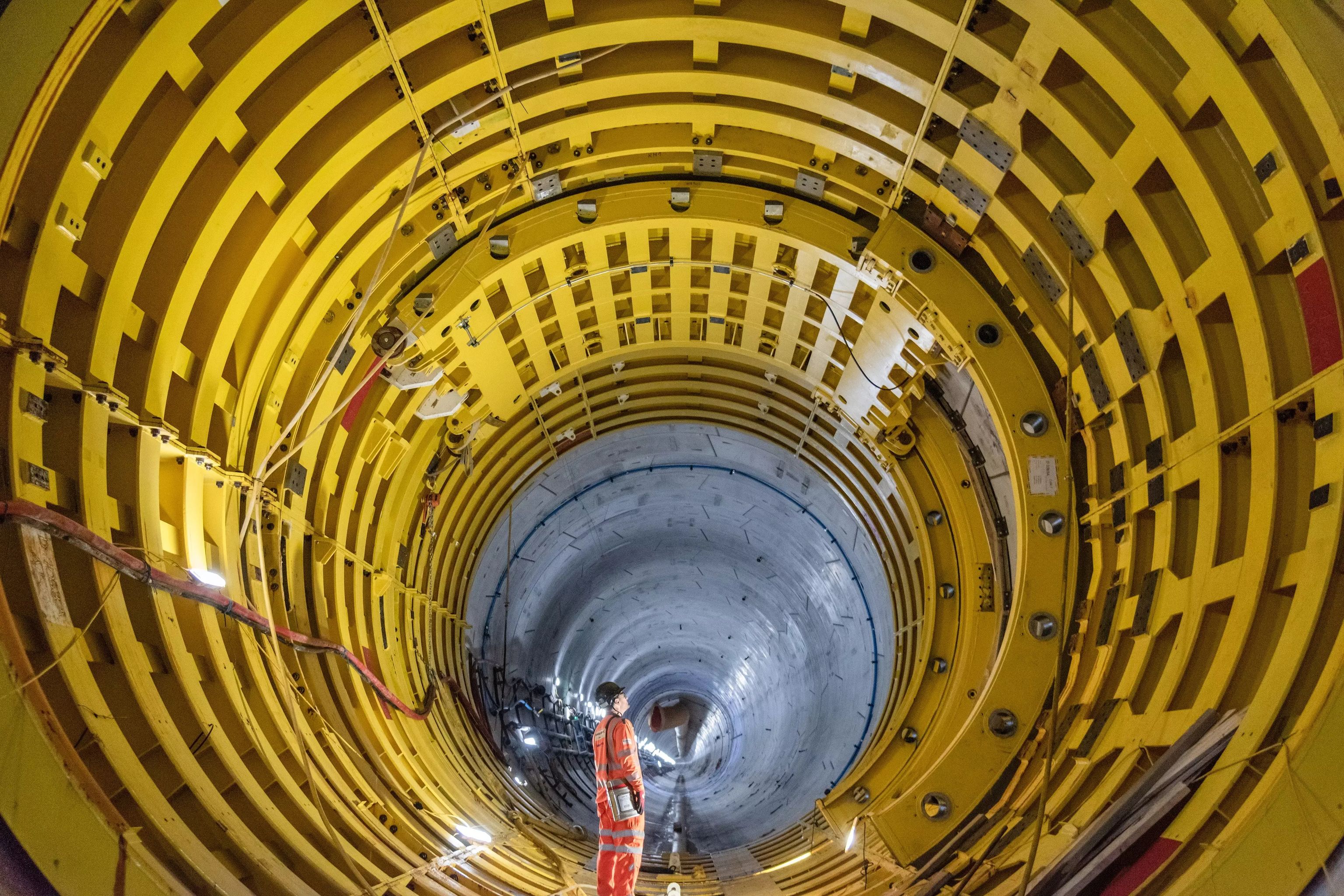 A worker stands in a large tunnel, framed by bright yellow metal rings, looking towards a gray concrete section.