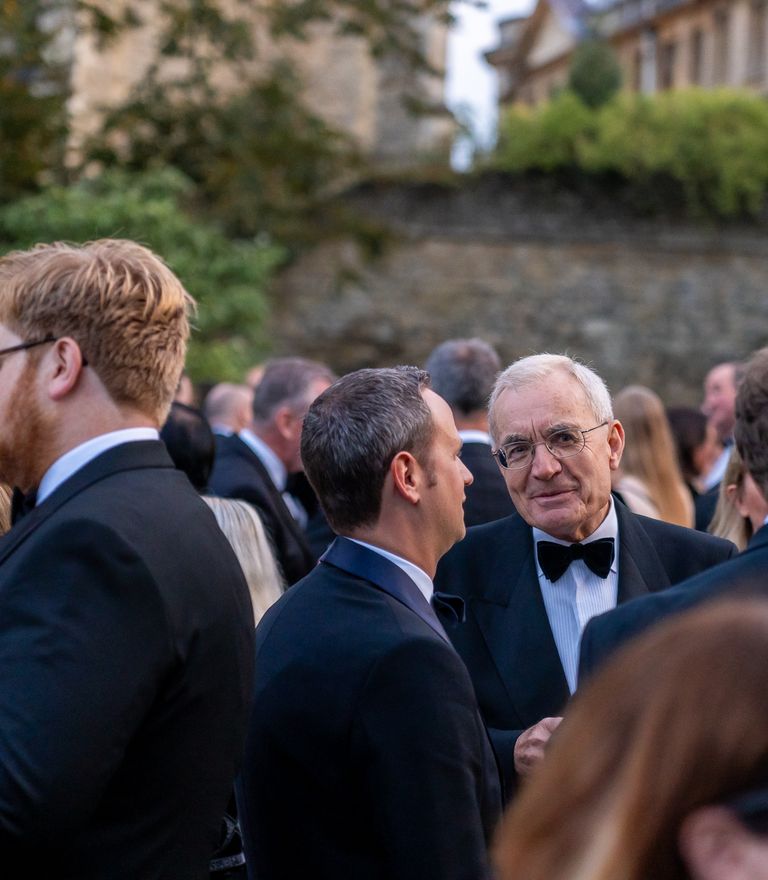 An outdoor gathering of people in formal wear, with an older man in a tuxedo smiling.