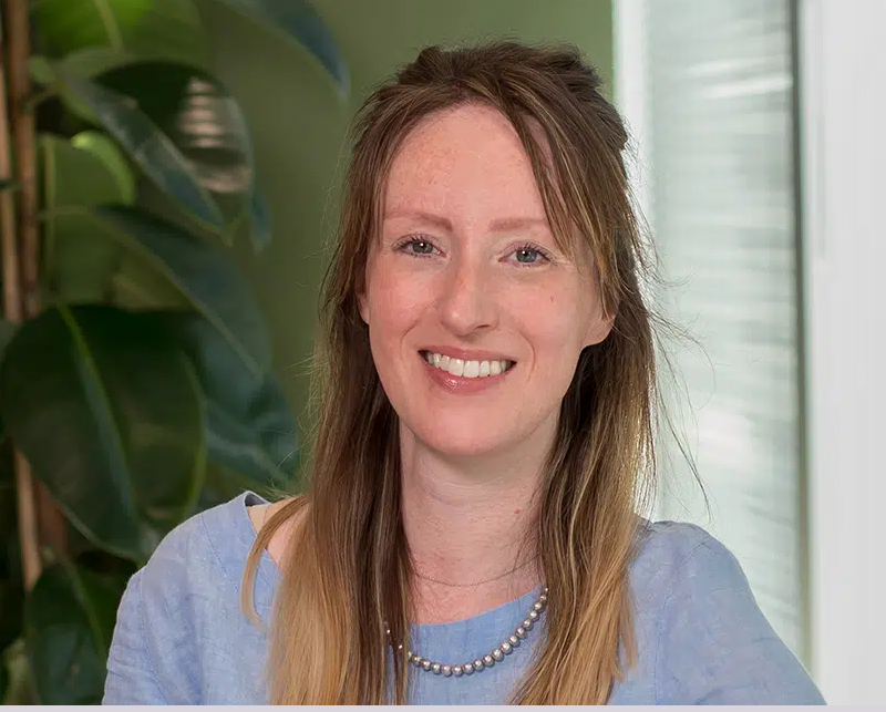 Smiling woman with long brown hair, wearing a light blue top and pearl necklace, with green plants in the background.
