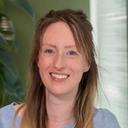 Smiling woman with long brown hair, wearing a light blue top and pearl necklace, with green plants in the background.