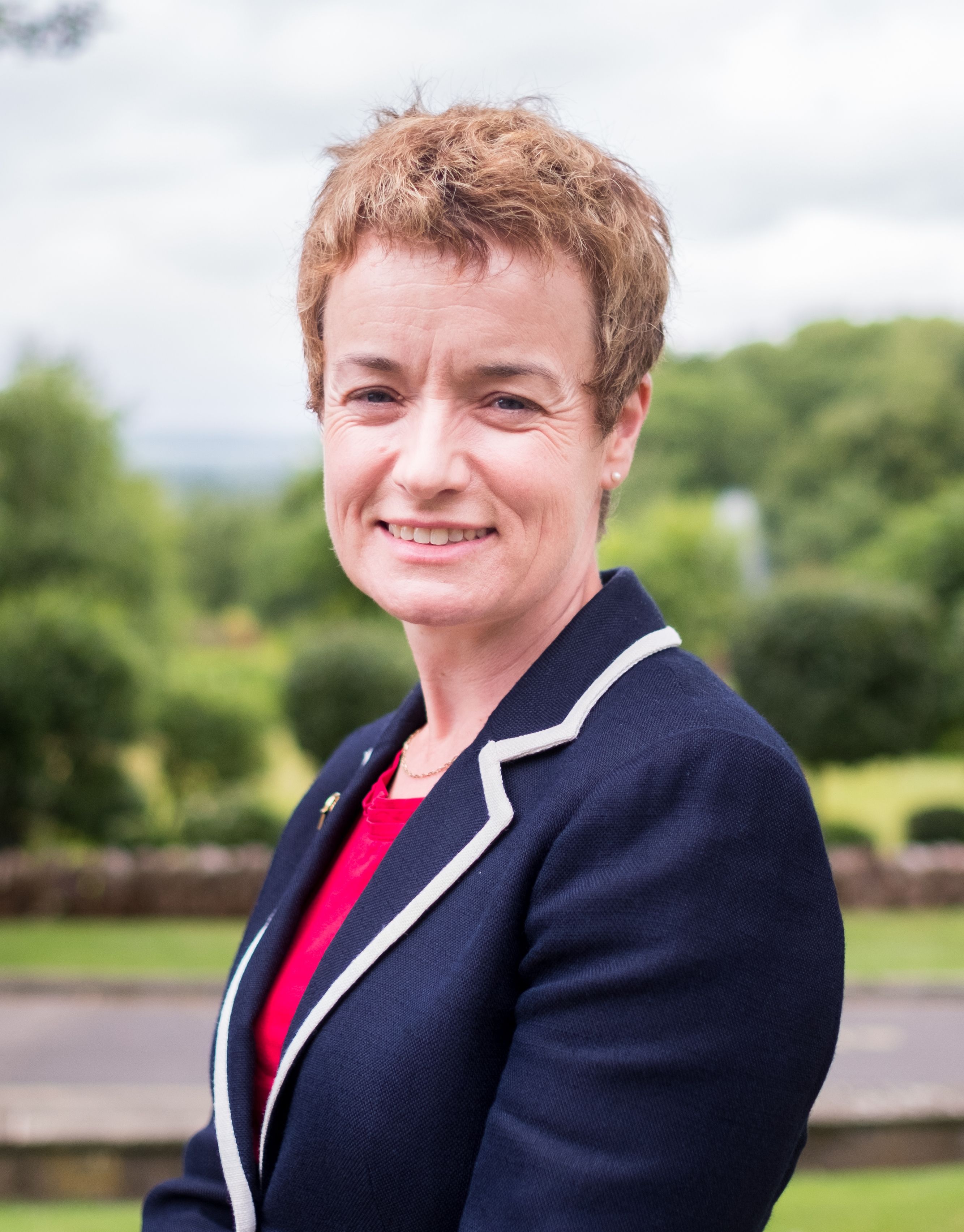 A smiling woman with short, curly brown hair wearing a blue blazer with white trim outdoors.