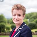 A smiling woman with short, curly brown hair wearing a blue blazer with white trim outdoors.