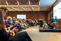 Audience at a conference listening to two speakers on stage, with a screen showing "Opening Address".