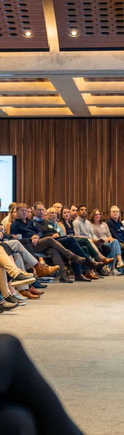 Audience at a conference listening to two speakers on stage, with a screen showing "Opening Address".