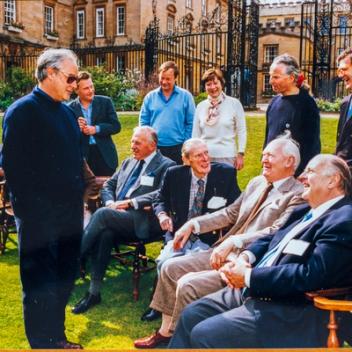 A group of men and a few women socialize on a sunny lawn in front of an ornate building with large gates.