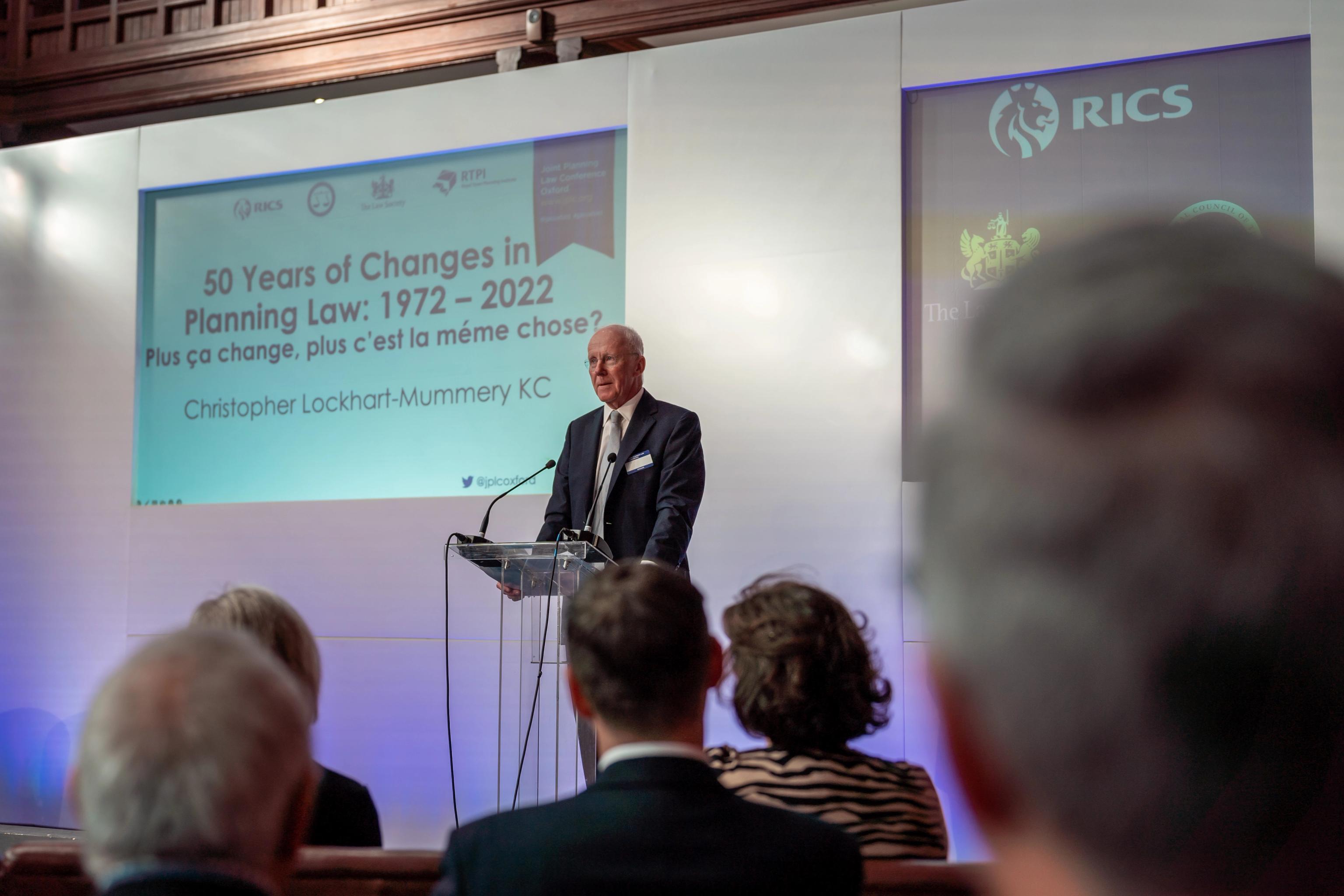 Christopher Lockhart-Mummery KC speaks at a podium before a screen displaying "50 Years of Changes in Planning Law: 1972 – 2022," observed by an audience.