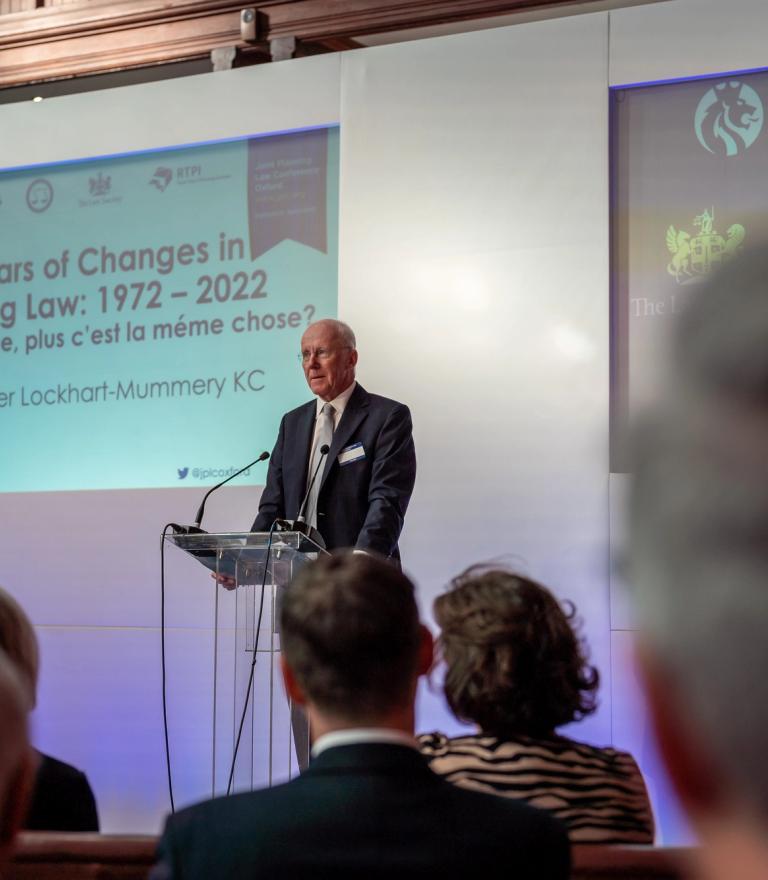 Christopher Lockhart-Mummery KC speaks at a podium before a screen displaying "50 Years of Changes in Planning Law: 1972 – 2022," observed by an audience.