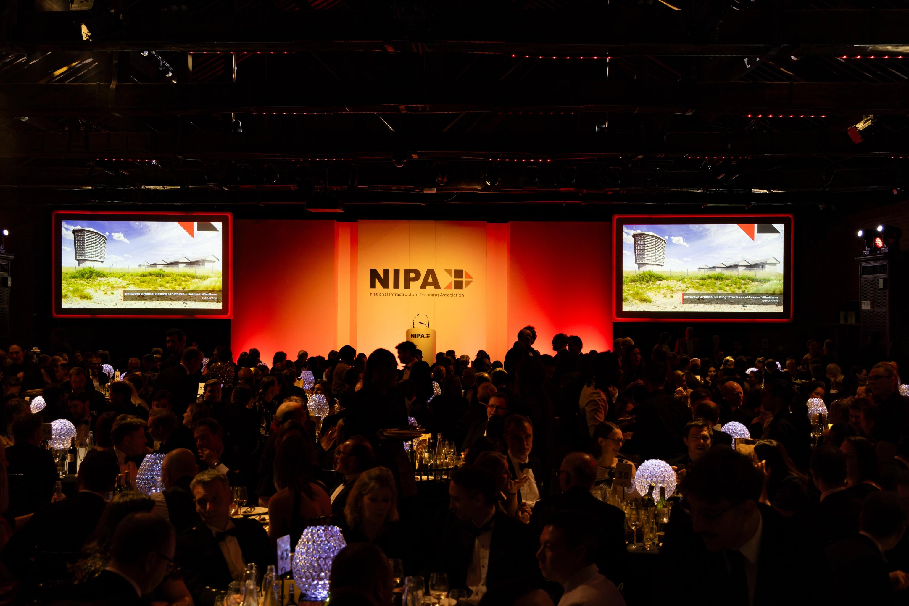 A large, dimly lit hall filled with people at tables, facing a stage with a prominent NIPA logo and screens showing a building design.