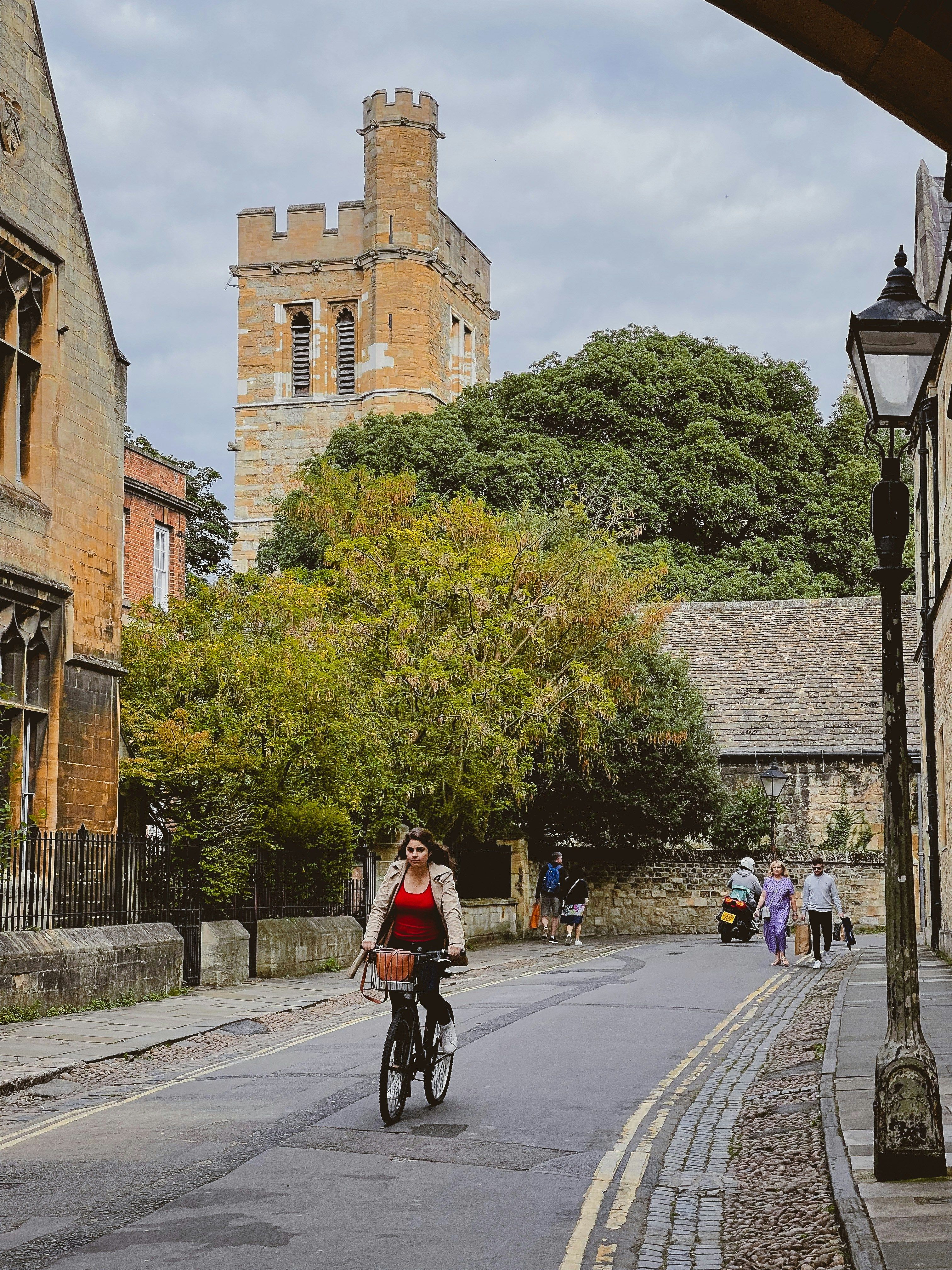 A woman rides a bicycle down a narrow, historic street with stone buildings, trees, and a tall tower under a cloudy sky.