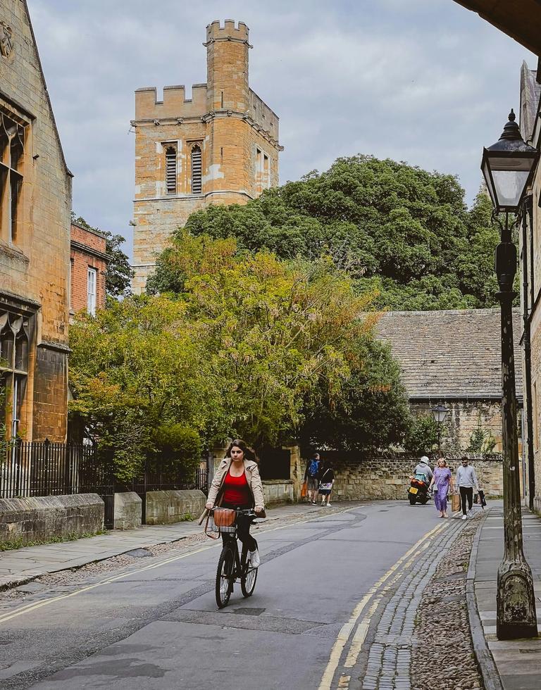 A woman rides a bicycle down a narrow, historic street with stone buildings, trees, and a tall tower under a cloudy sky.