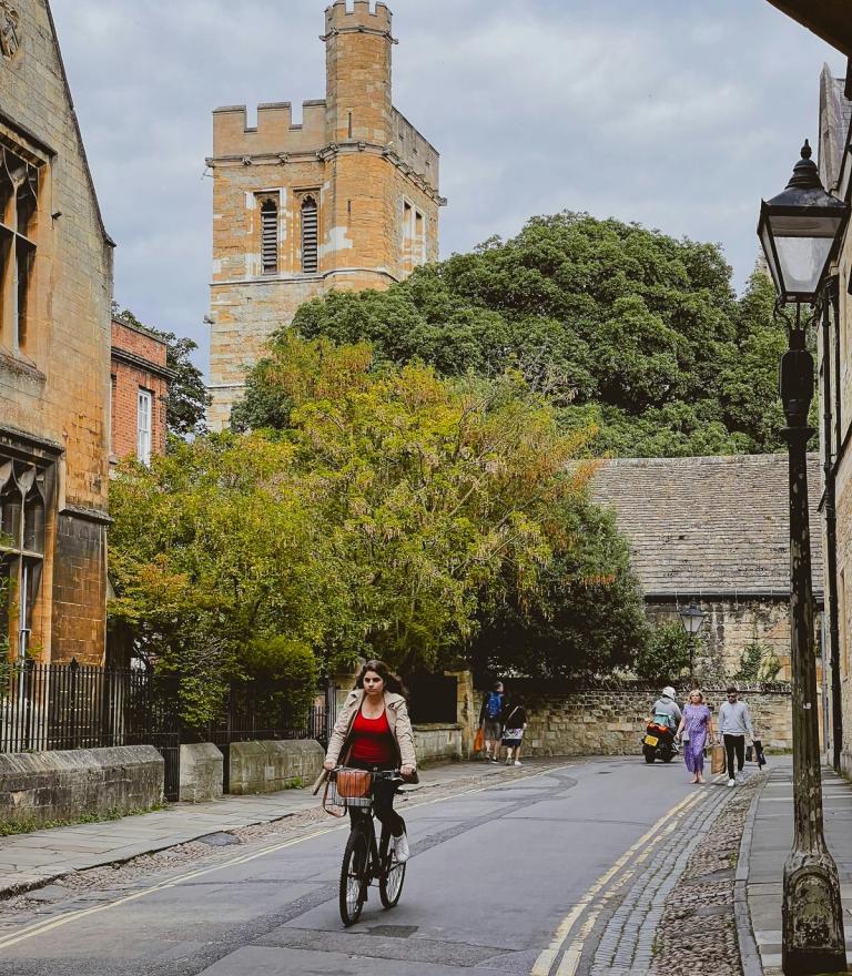 A young woman on a bicycle rides down a historic street with a stone tower in the background.