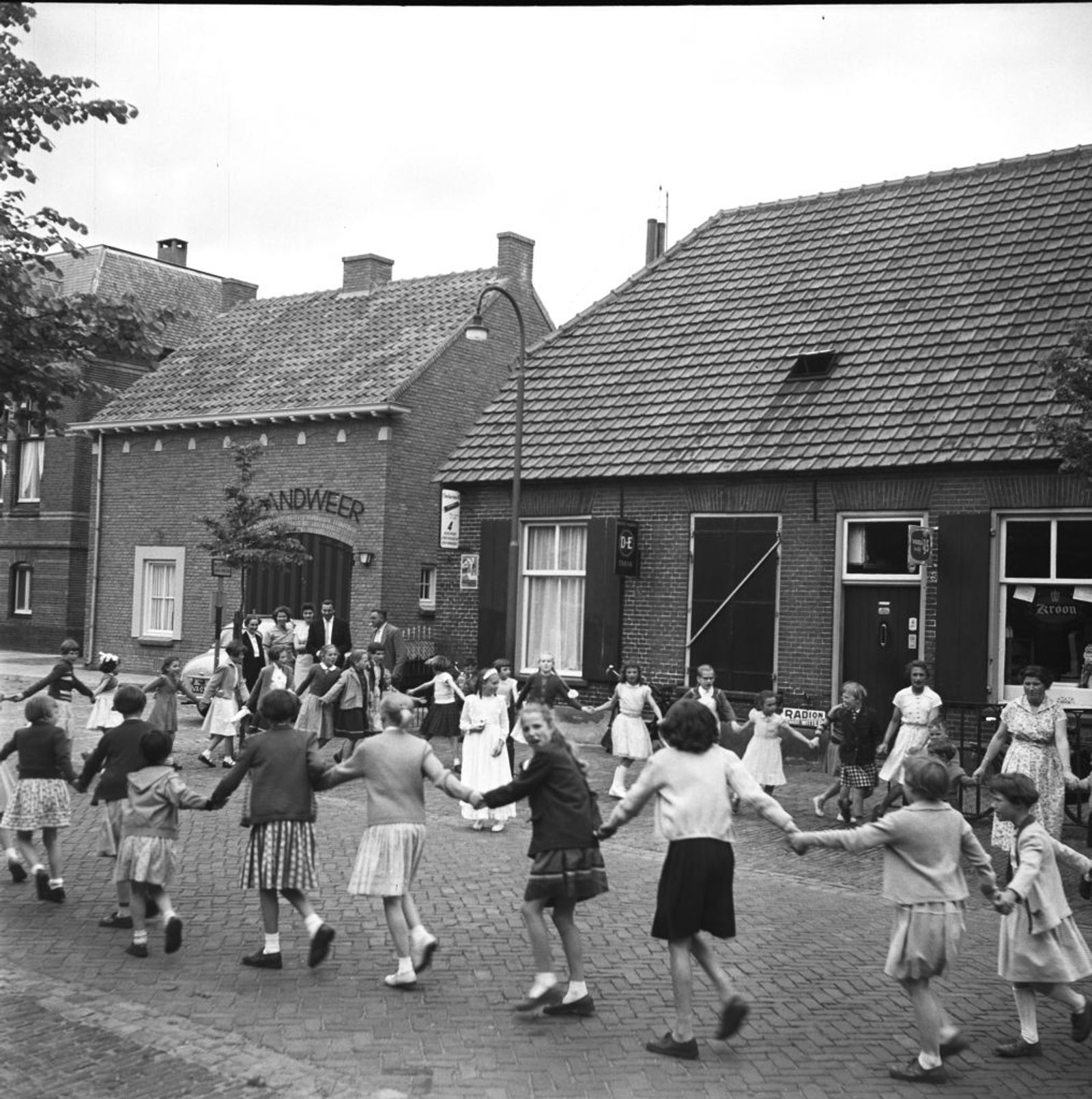 Pinksterblom (pinksterbruidje) Lia Wouters. De kinderen dansen in een kring om haar heen. Naast de Kroonwinkel van de familie Wouters de brandweerkazerne. (Zie ook foto: 1634-011984)