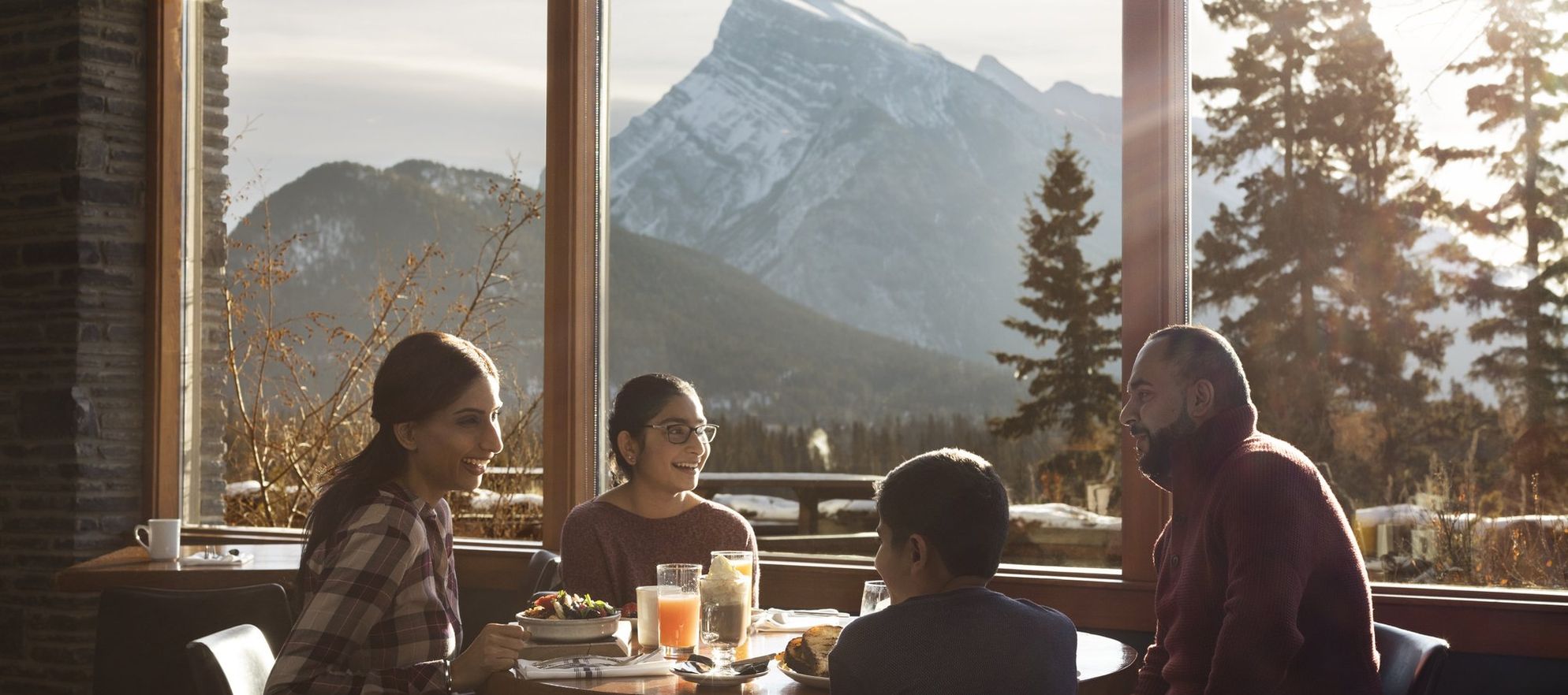 A family of four enjoying a meal with Rundle Mountain and snow visible in the window behind them