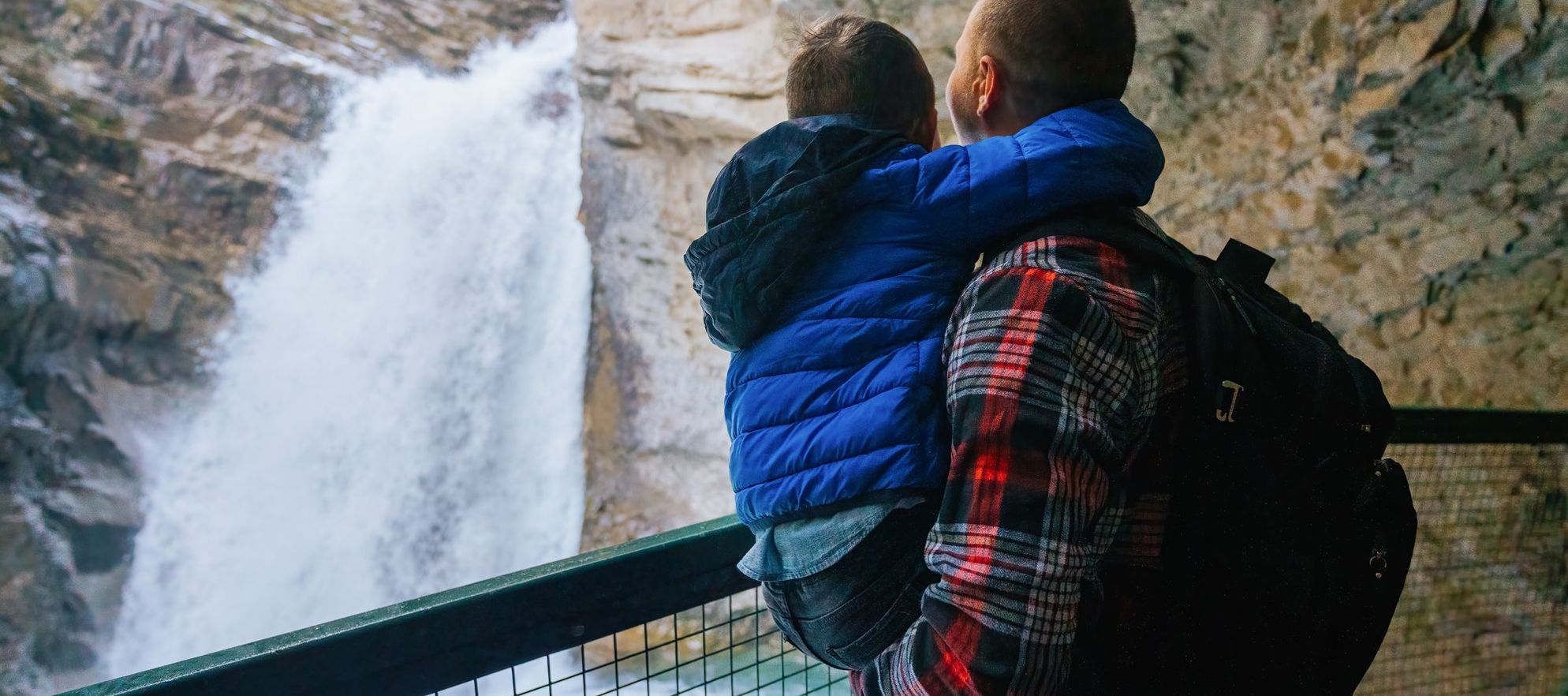 A father wearing a red flannel shirt holds his son in a bright blue jacket as they look at a rushing waterfall from behind the safety of a guardrail