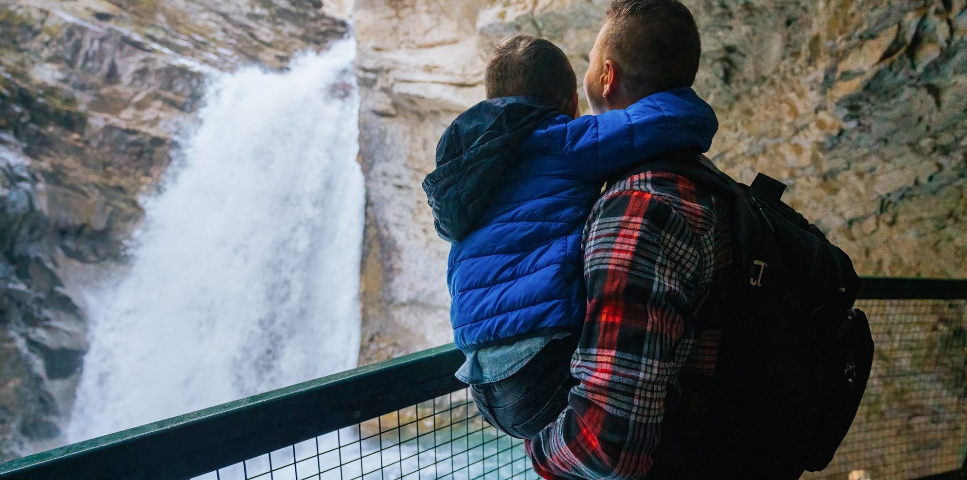 A father wearing a red flannel shirt holds his son in a bright blue jacket as they look at a rushing waterfall from behind the safety of a guardrail