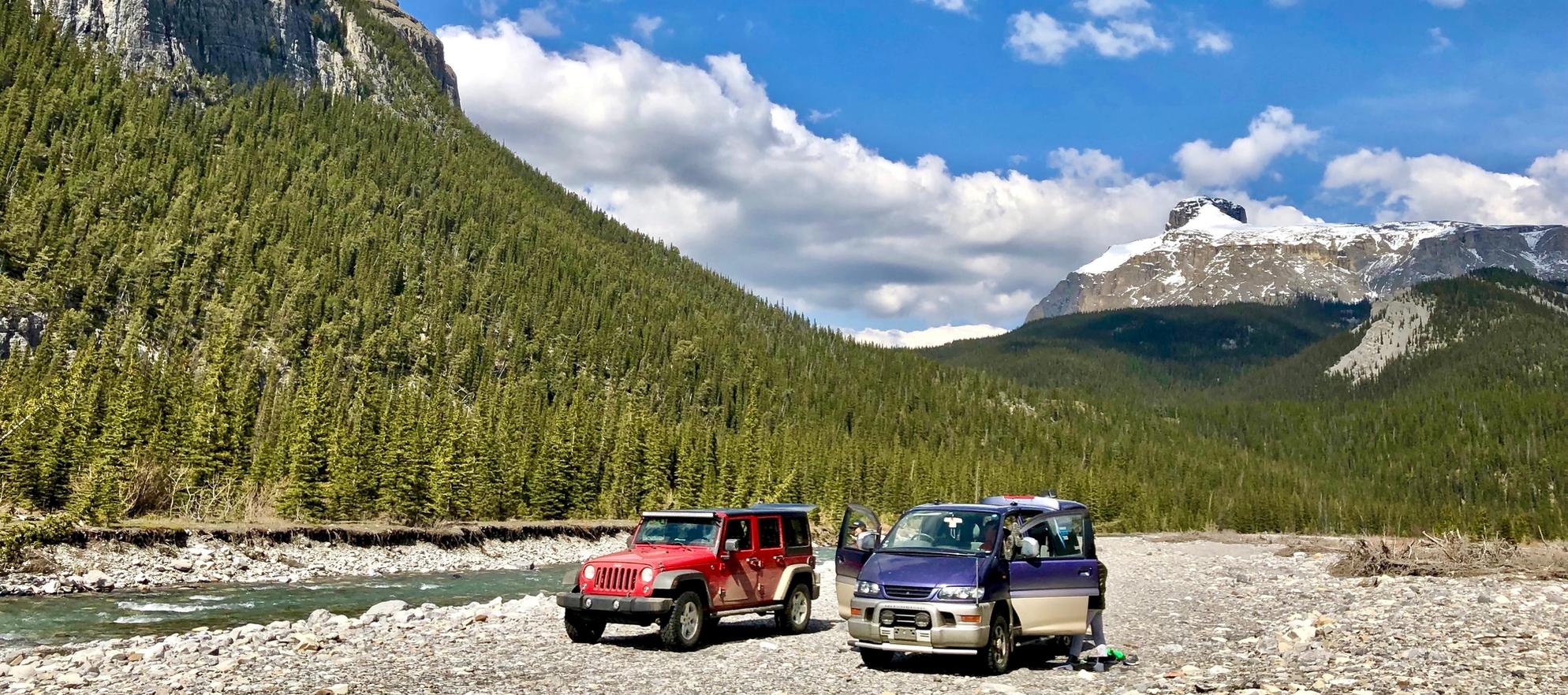 A Jeep and a van parked off-road with mountains and a river in the background
