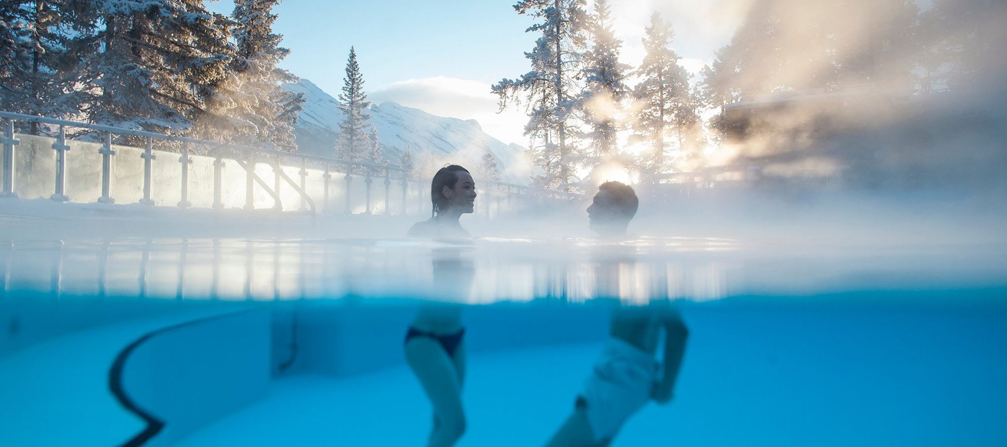 Two people in the Banff Upper Hot Springs in Banff National Park with sunlight pouring through the trees behind them.