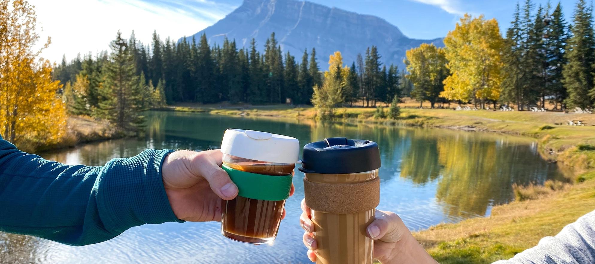 Two coffee cups "cheers-ing" at Cascade Ponds. Rundle Mountain and Fall colours in the background