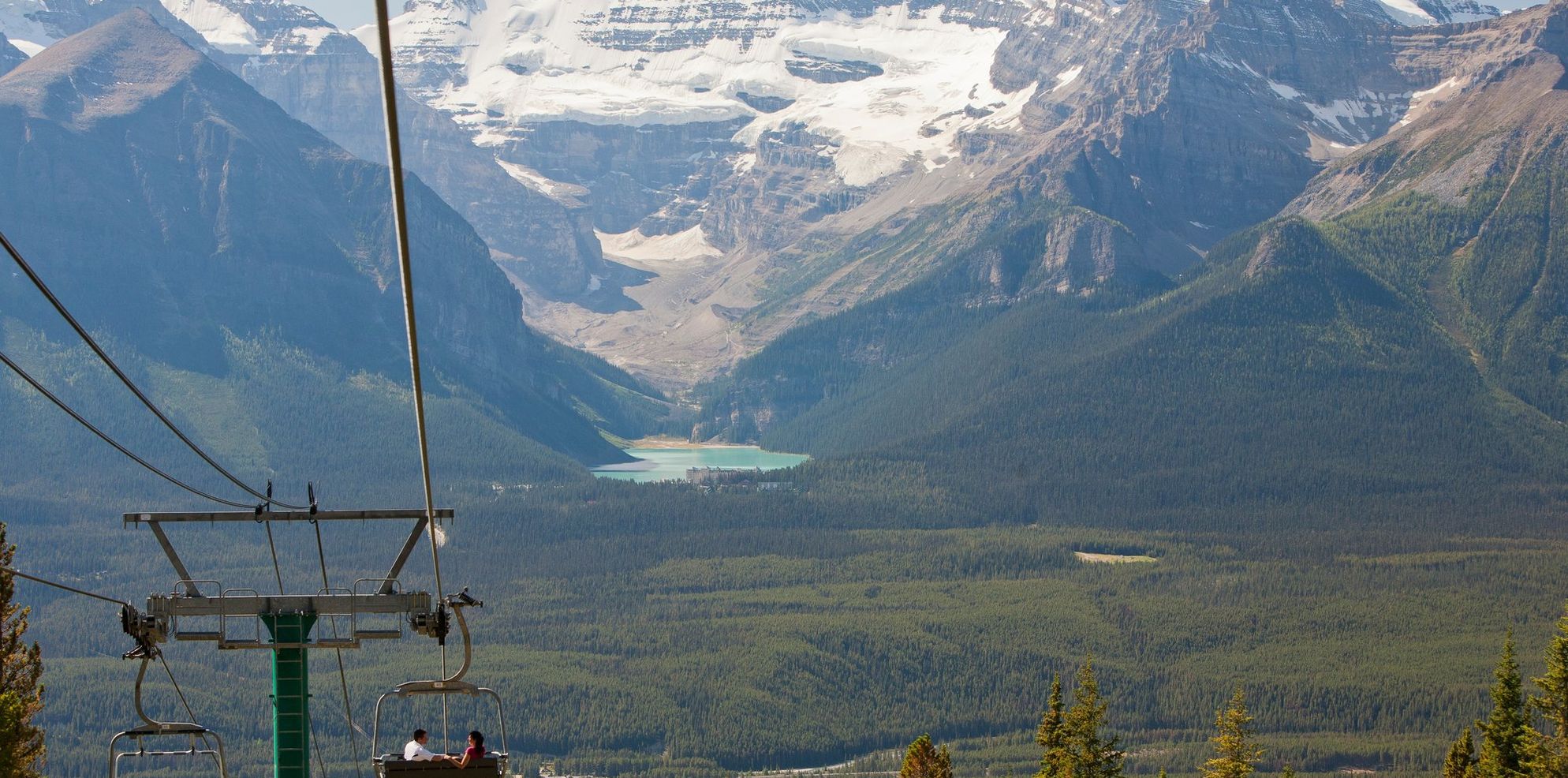 A couple travels down a mountain in an open chairlift while taking in the view of Lake Louise and Victoria Glacier in the distance