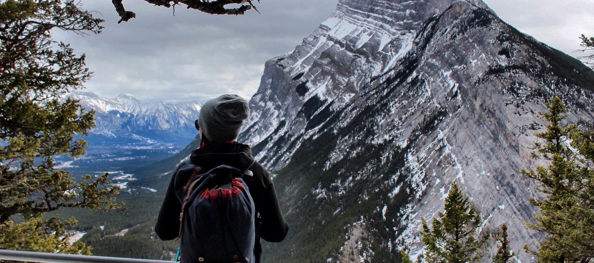 A woman stands on Tunnel Mountain looking towards Mountain Rundle surrounded by trees in Banff National Park.