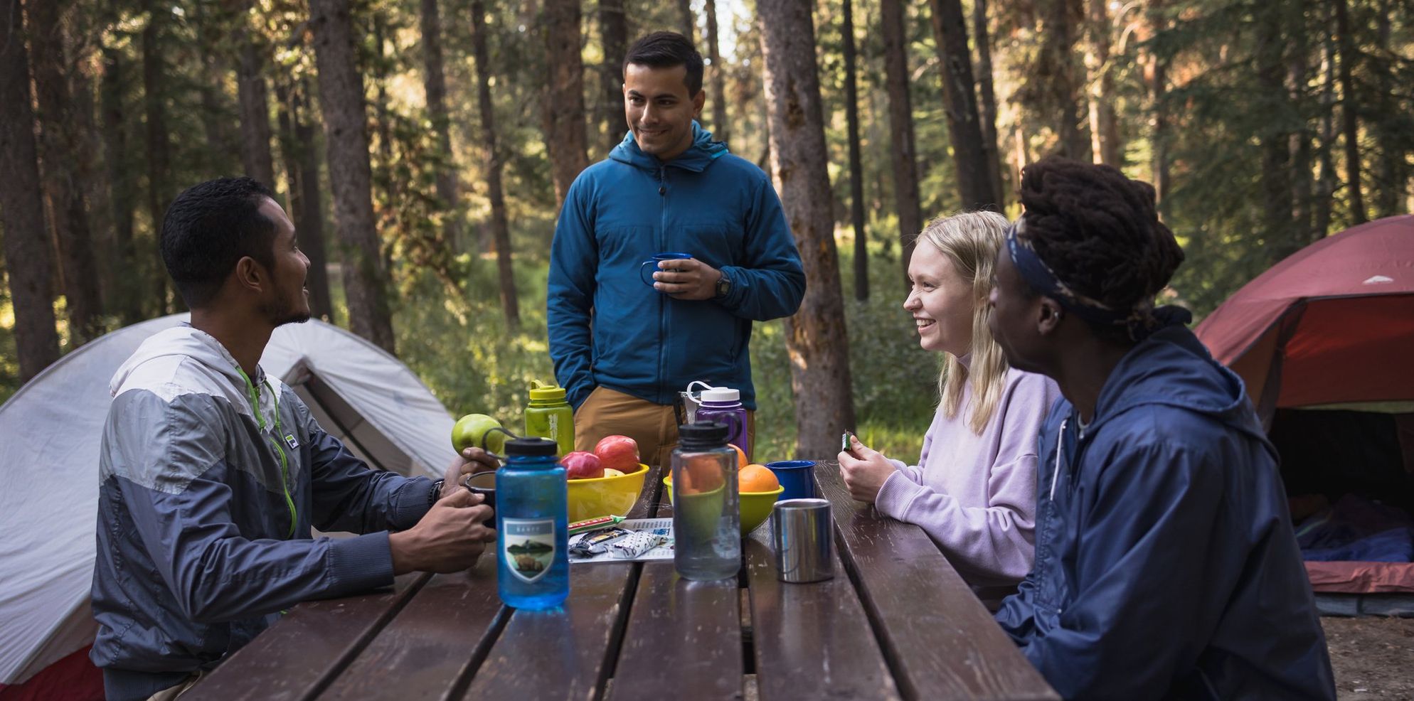 A group of campers sit at a picnic table with snacks and water - two tents can be seen in the background
