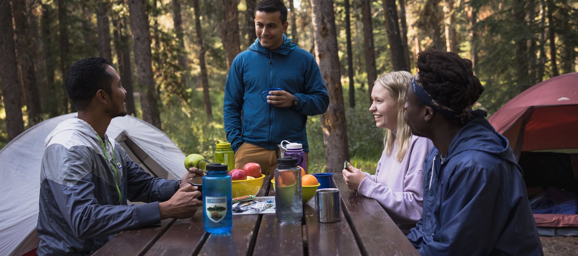 A group of campers sit at a picnic table with snacks and water - two tents can be seen in the background