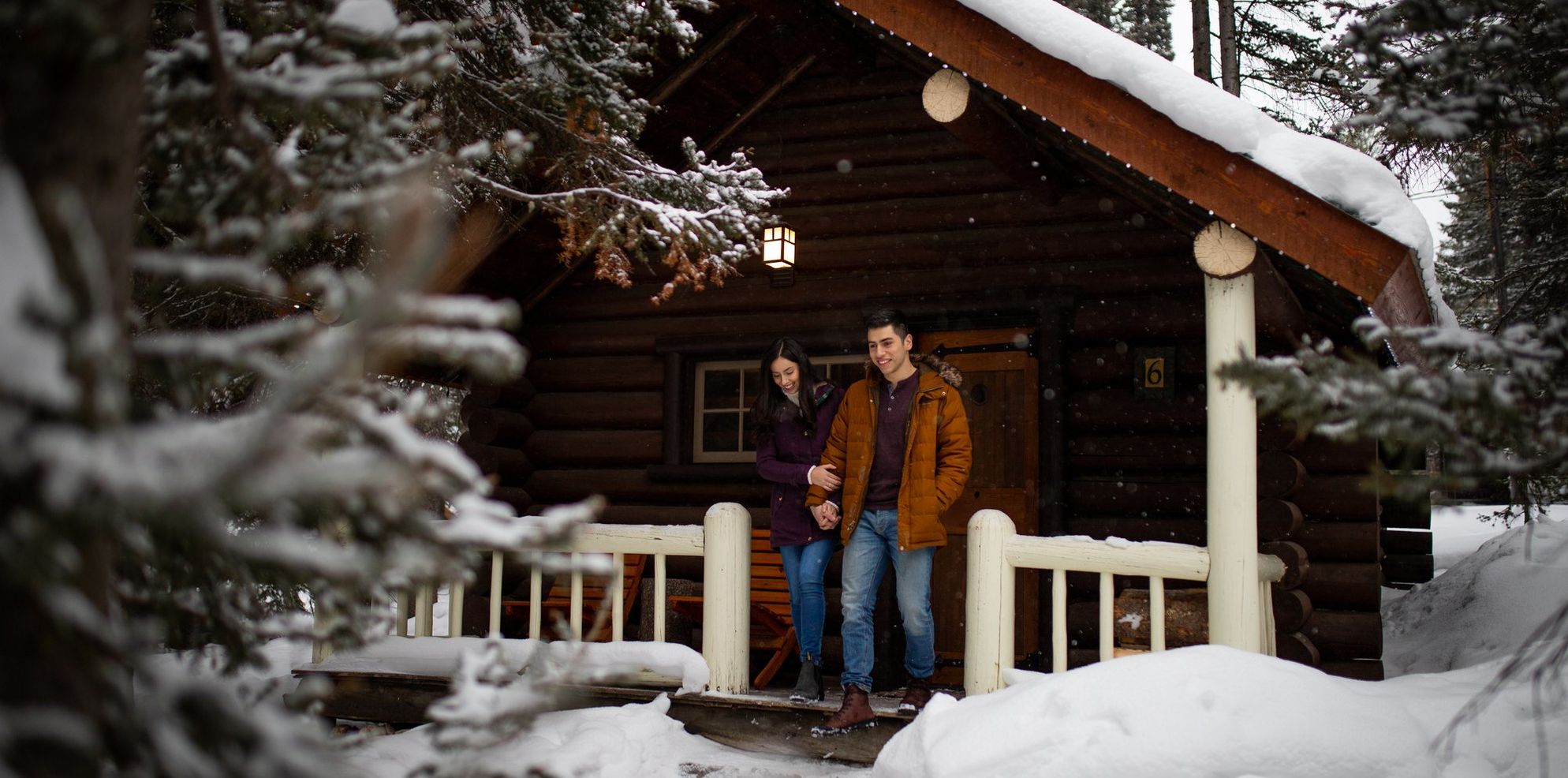 A couple in front of a cozy winter cabin surrounded by snowy trees in Banff National Park.