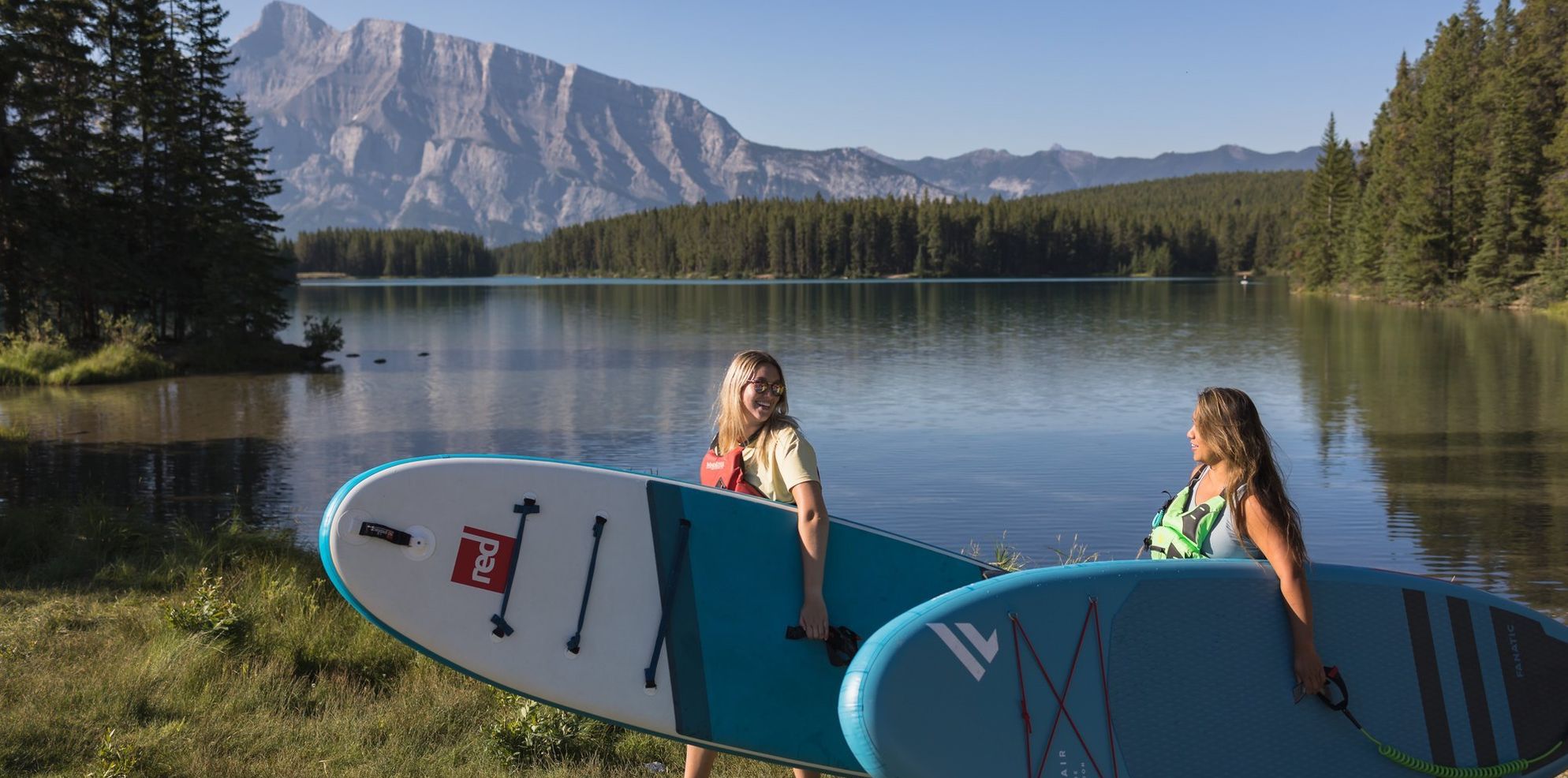Two friends walk with paddleboards in hand to a stunning turquoise lake surrounded by mountains