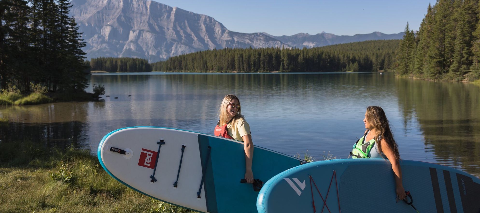 Two friends walk with paddleboards in hand to a stunning turquoise lake surrounded by mountains