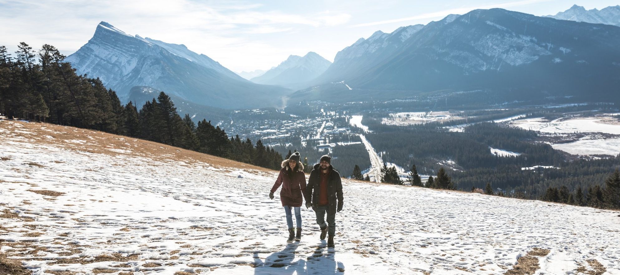 A couple hiking on a hill with grass peeking through the snow and an aerial view of the town and the mountains behind them