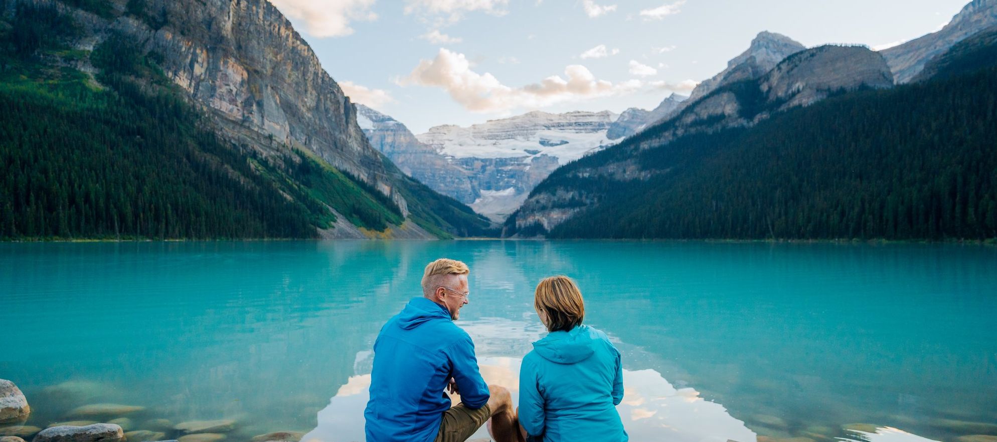 A mature couple sits on the rocks at the edge of a vast turquoise blue lake with a glacier and large mountains in view at the end of the lake