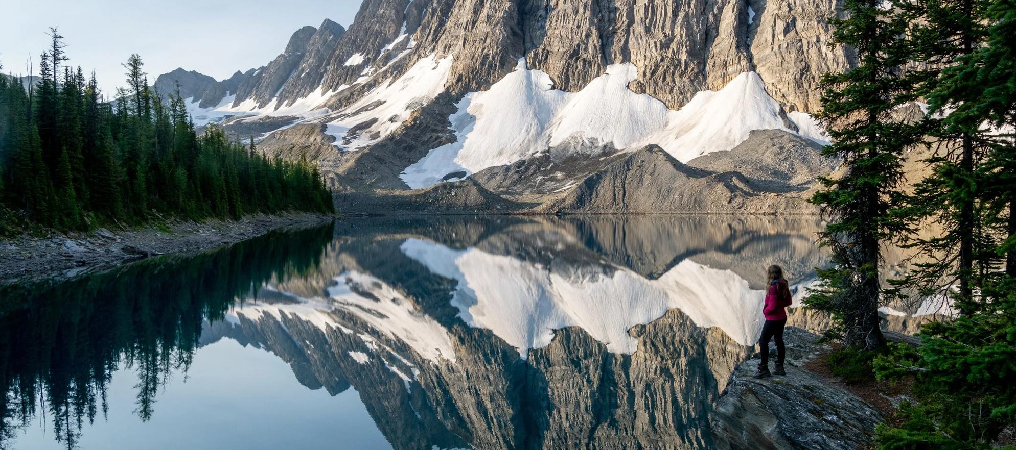 a woman in a red jacket gazing over a lake at sunrise with mountains reflecting in the water