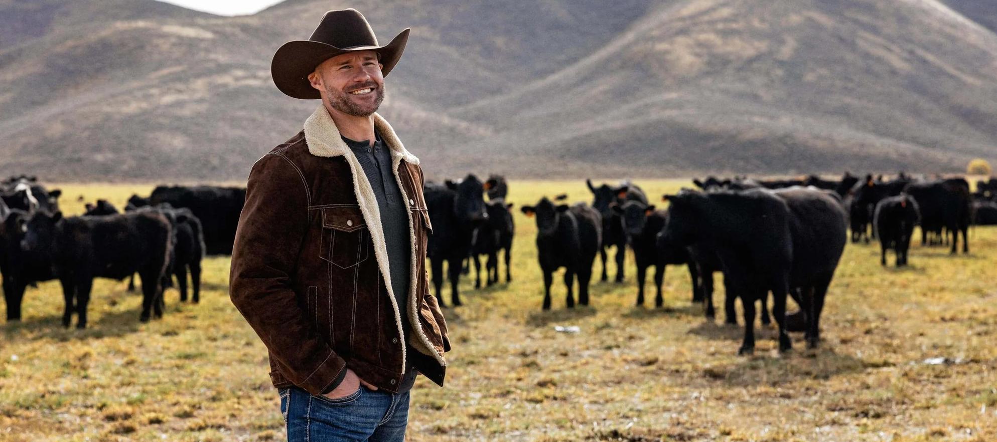 A man in a cowboy hat standing in a field with a herd of black bows in the background