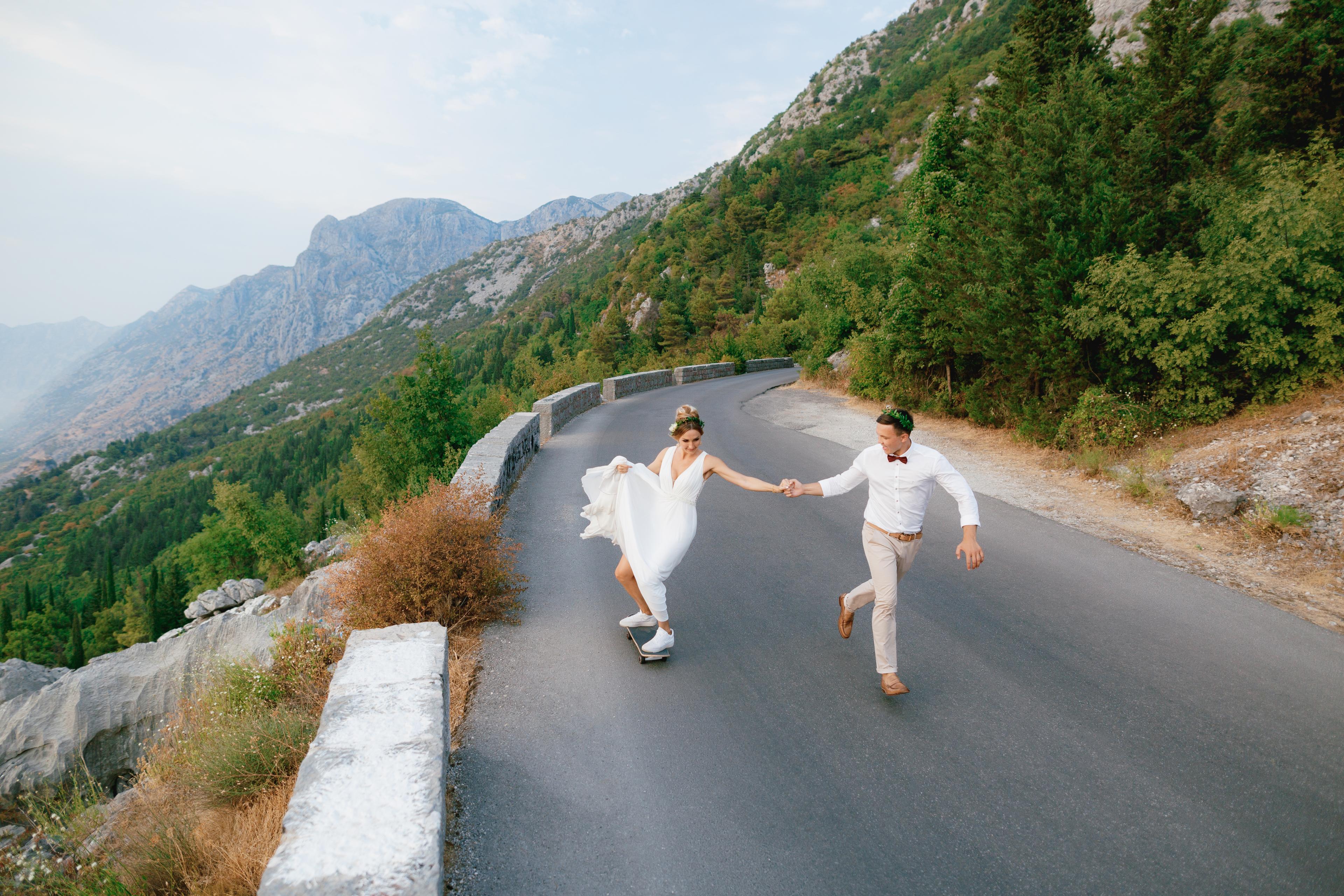 A bride and groom joyfully skateboarding on a winding mountain road, surrounded by lush greenery and mountains.