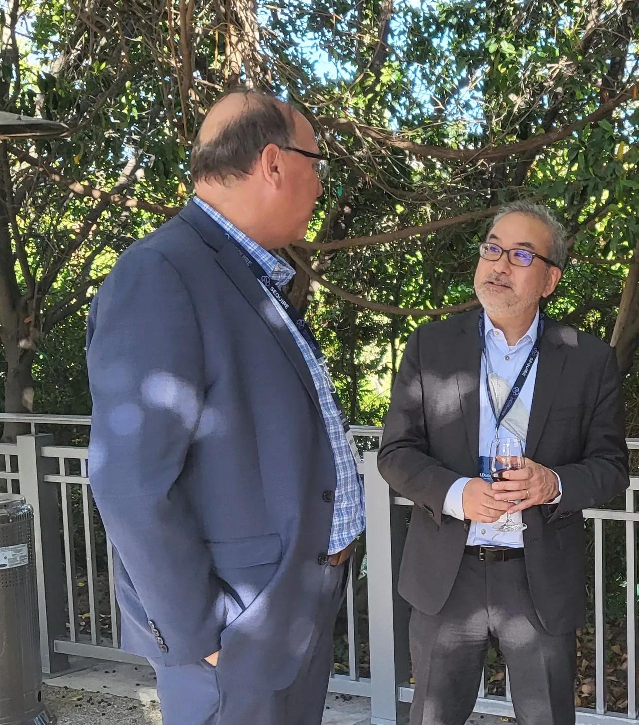 Two men in suits, GIX's Senior Listings Advisor Marc Iyeki and Board Member Rob Wotczak, talking outdoors. Wotczak is turned away from the camera and Iyeki is leaning on a railing.