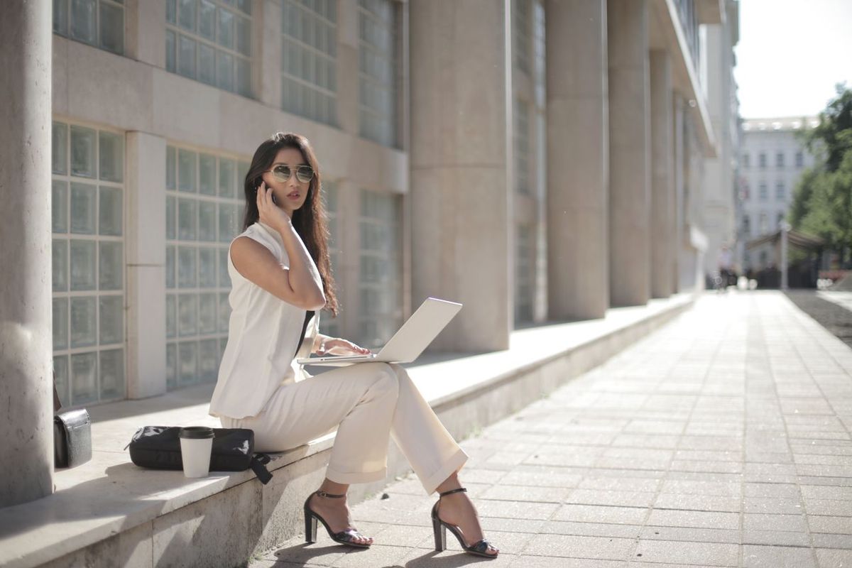 Person seated outdoors with a laptop, talking on a smartphone beside an office building.