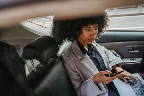 Woman multitasking with phone and laptop in car backseat, balancing work and travel.