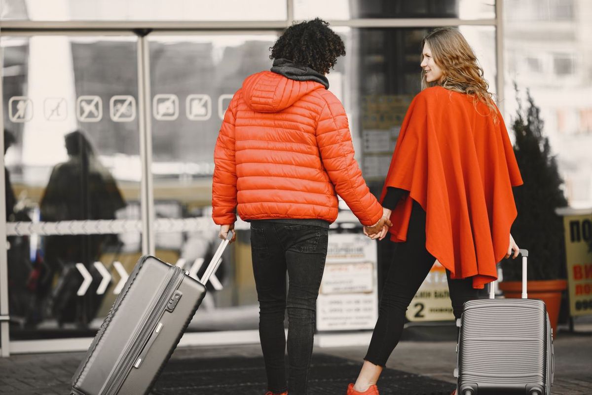 Two travelers pulling suitcases and holding hands outside a building entrance.