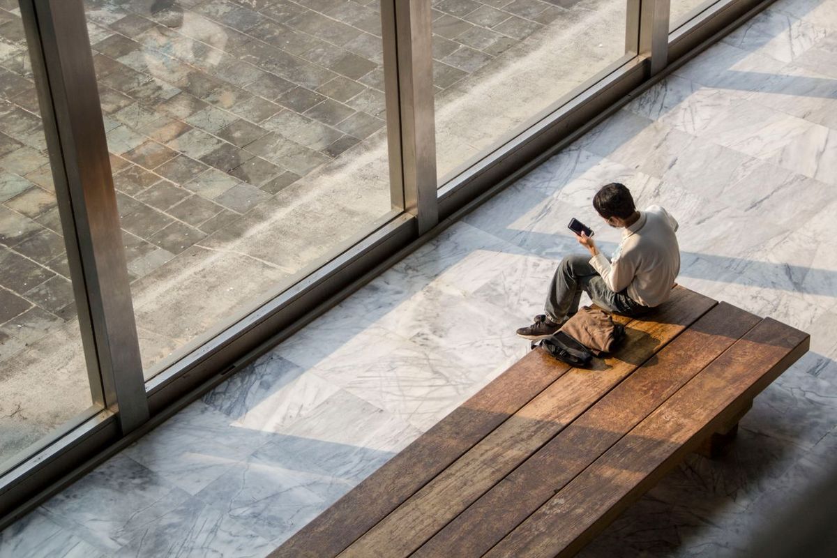 Person sitting on a bench by large windows while looking at a smartphone.