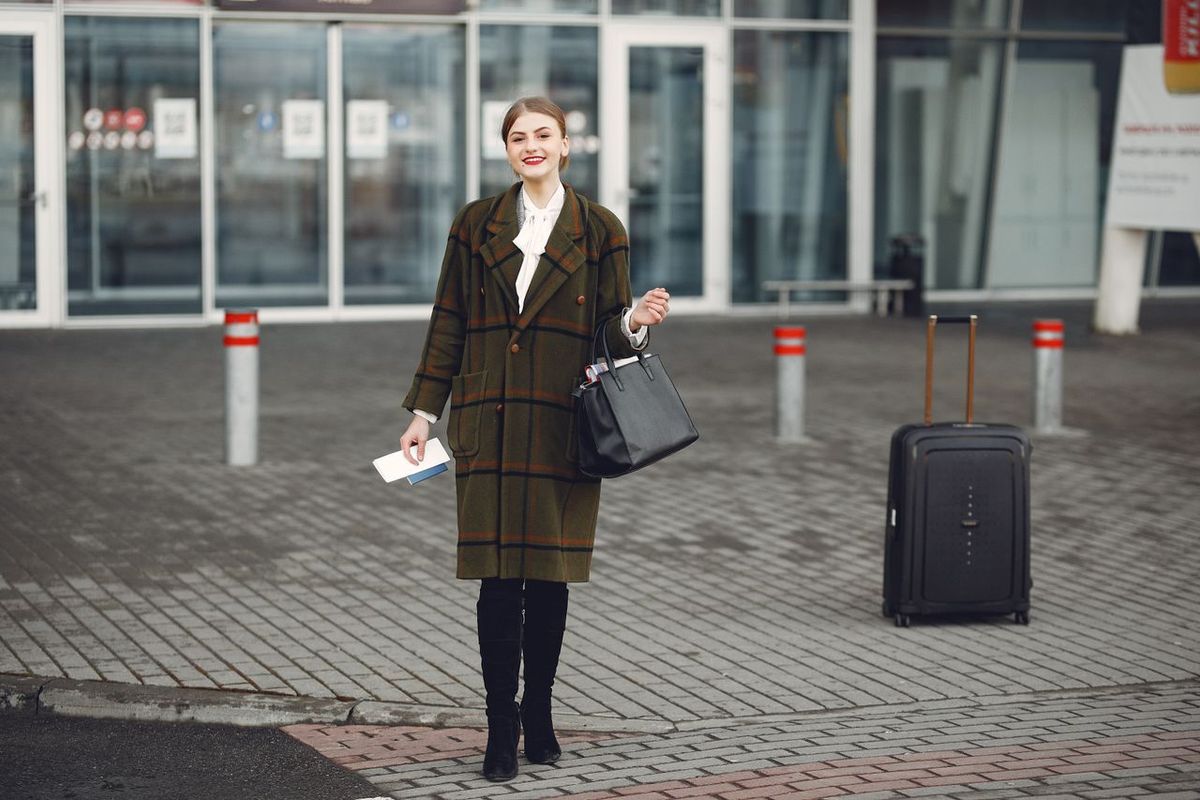 Person standing outside a building holding travel documents, with a suitcase nearby.