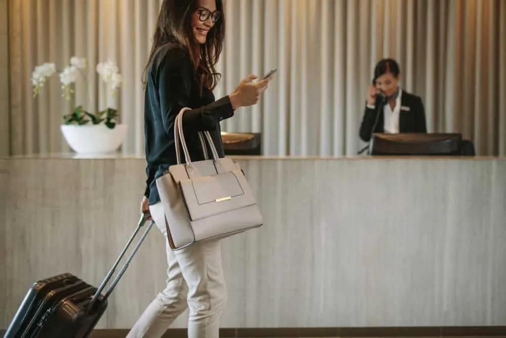 Woman using mobile phone and pulling her suitcase in a hotel lobby. Female business traveler walking in hotel hallway.